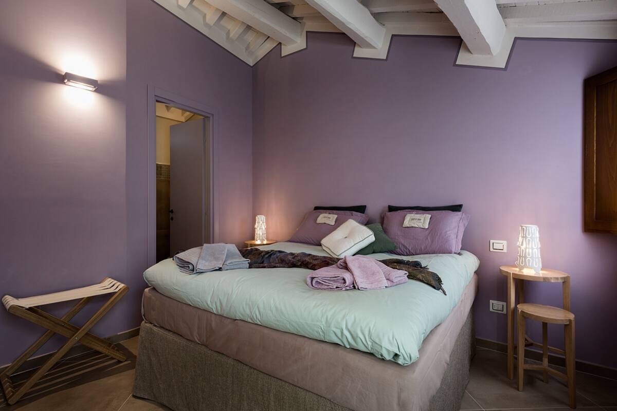 Een slaapkamer met een bed, kussens, een nachtkastje, een hanglamp en een houten kruk.