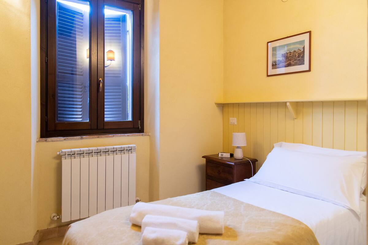 Een hotelkamer met een bed, een nachtkastje, een ingelijste foto aan de muur en een raam met jaloezieën.