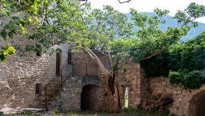 An old stone building with a staircase leading to a balcony, surrounded by greenery and a mountainous backdrop.