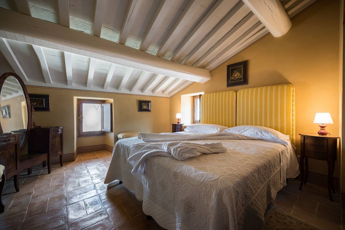 Een slaapkamer met een groot bed, een houten dressoir, een spiegel en een raam met gordijnen.