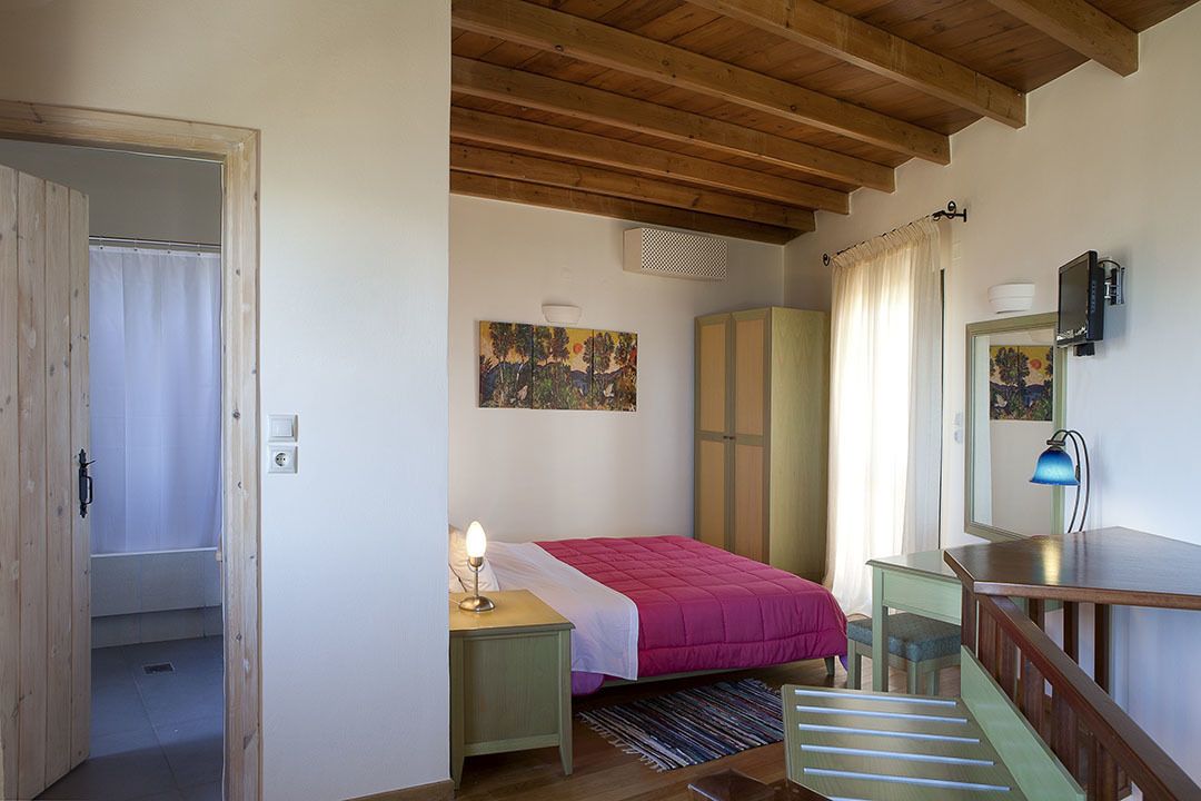 A bedroom with a wooden ceiling, a bed with a pink bedspread, a nightstand with a lamp, a wardrobe, a desk with a chair, and a small staircase.