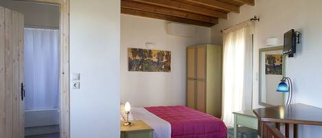 A bedroom with a wooden ceiling, a bed with a pink bedspread, a nightstand with a lamp, a wardrobe, a desk with a chair, and a small staircase.