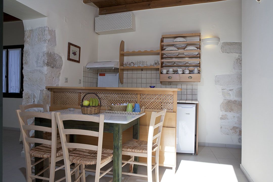 A compact kitchen with a wooden table and chairs, a small refrigerator, and a shelf with dishes and glasses.