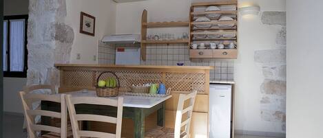 A compact kitchen with a wooden table and chairs, a small refrigerator, and a shelf with dishes and glasses.