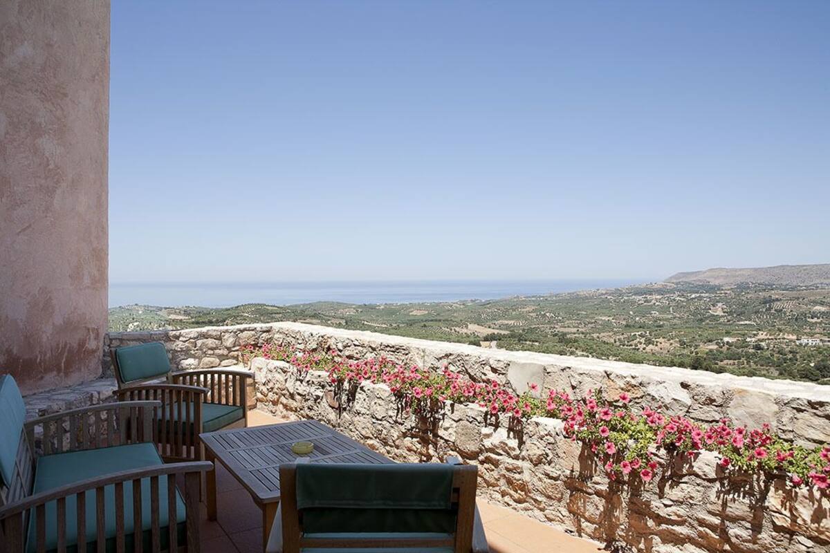 Terraço com mesa e cadeiras, com vista para uma paisagem florida em tons de rosa e uma paisagem distante.