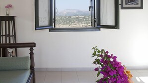 A room with a wooden ceiling, a large open window with curtains, a framed picture on the wall, a green sofa, a round table with a vase of flowers, and a book. - Kastellos Village - Central Complex (Apokoronas)
