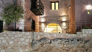 A stone pool with a clear blue edge, a stone wall, and a building with a balcony and a lit-up archway. - Kastellos Village - Central Complex (Apokoronas)