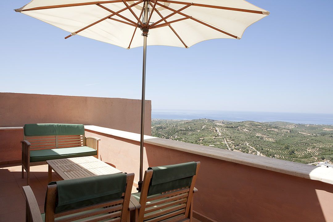 A balcony with a wooden table and chairs, a large umbrella, and a view of a hilly landscape.