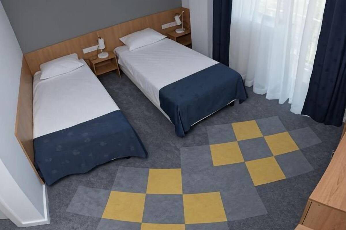 A hotel room with two beds, a wooden headboard, a bedside table with a lamp, a checkered carpet, and a window with curtains.