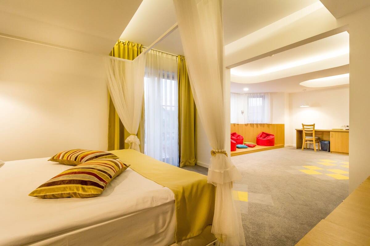 A hotel room with a bed, a desk, and a chair. There is a curtain and a window with curtains.