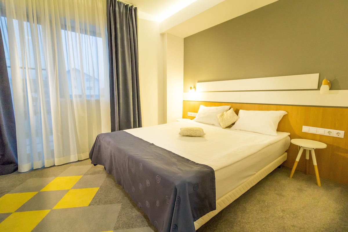 A hotel room with a bed, a bedside table, a window with curtains, and a wall-mounted light fixture.