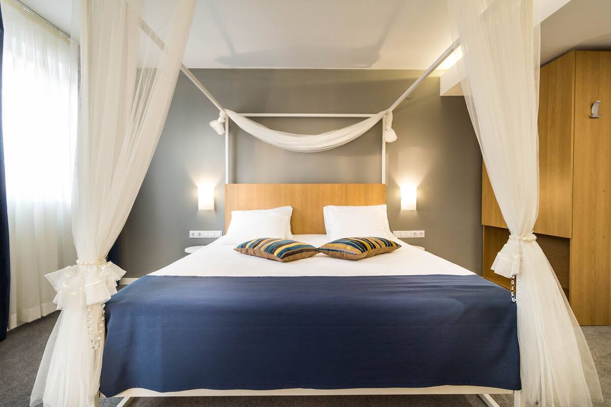 A bed with a blue bedspread and two pillows, a wooden headboard, and white curtains.