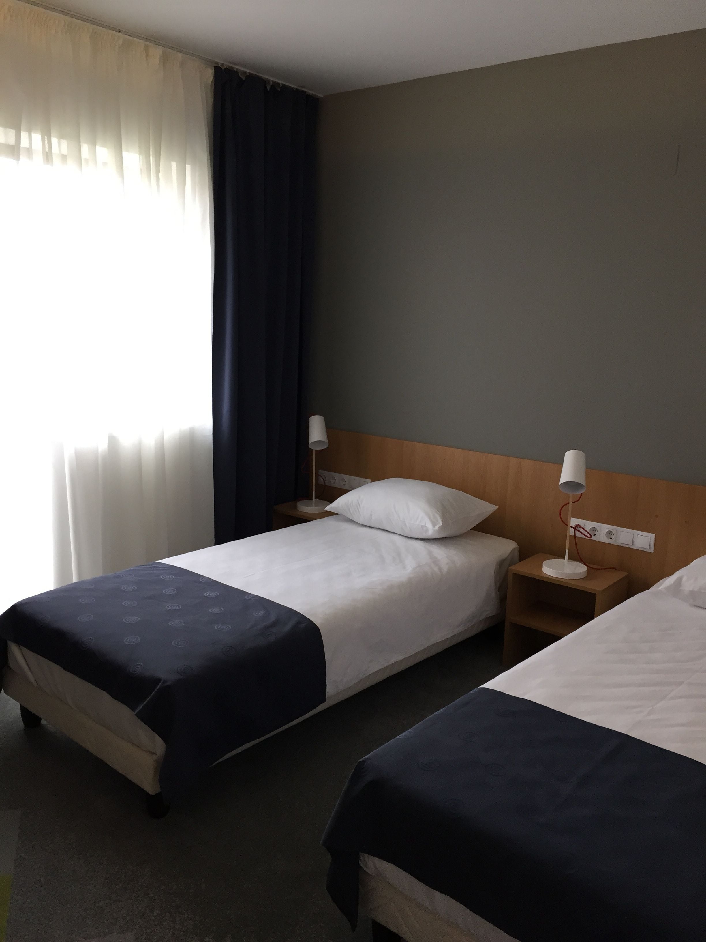 A hotel room with two beds, a wooden headboard, a bedside table with a lamp, and a window with curtains.