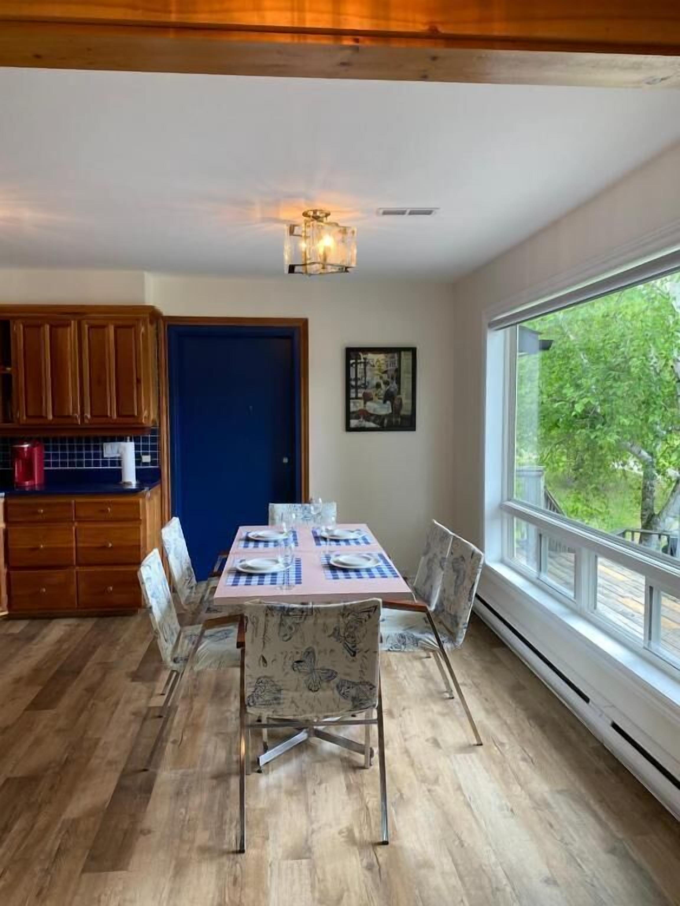 A dining area with a table set for four, wooden cabinets, a blue door, and a window with a view of trees.