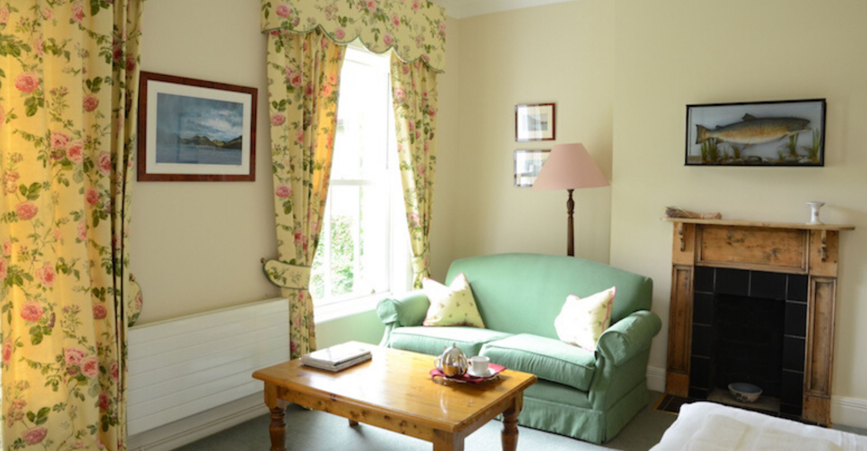 A room with a green sofa, a wooden table with a book and a teapot, floral curtains, a fireplace, and framed pictures on the wall.