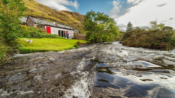 A stone building with a red door, a grassy area with a dog, and a flowing river. - Delphi Lodge & Estate (Leenane)