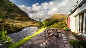 A riverside patio with a wooden table and benches, a dog, and a building with a red door. - Delphi Lodge & Estate (Leenane)