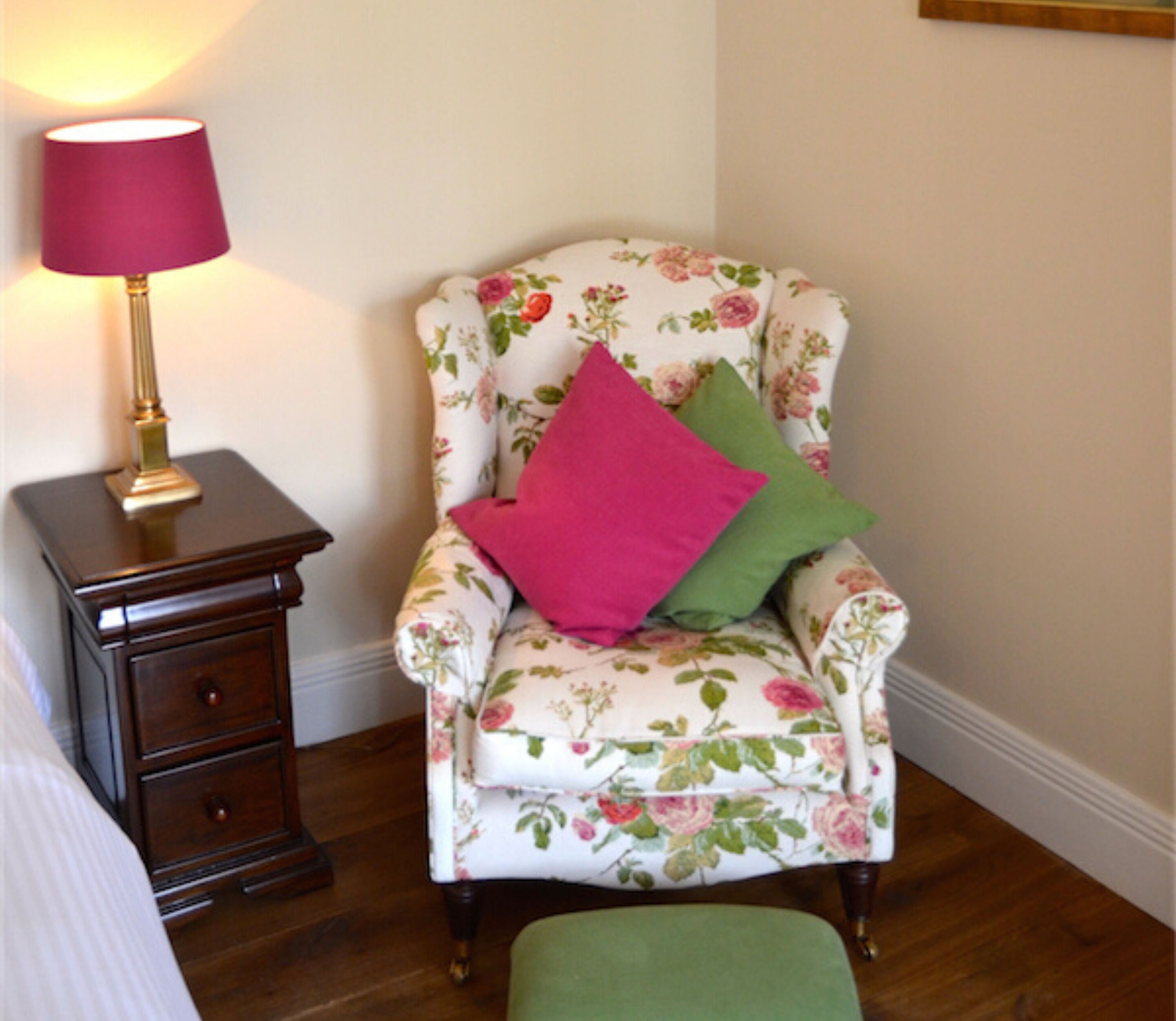A floral armchair with two pillows, a small wooden nightstand with a lamp, and a green ottoman.