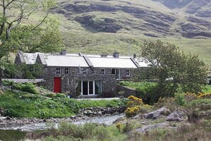 Ein Steinhaus mit roter Tür, eingebettet in eine üppige, grüne Landschaft mit einem Bach im Vordergrund.