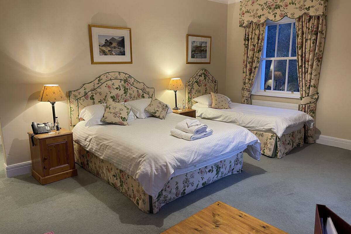 Ein Hotelzimmer mit zwei Betten, blumengemusterten Kopfteilen, Nachttischlampen, einem Holznachttisch, einem Holztisch und einem Fenster mit floralen Vorhängen.