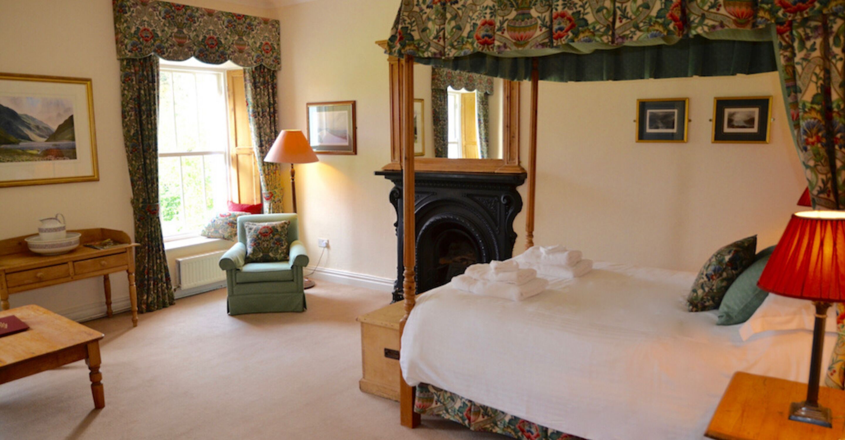 A bedroom with a four-poster bed, a fireplace, a desk with a lamp, and a chair.
