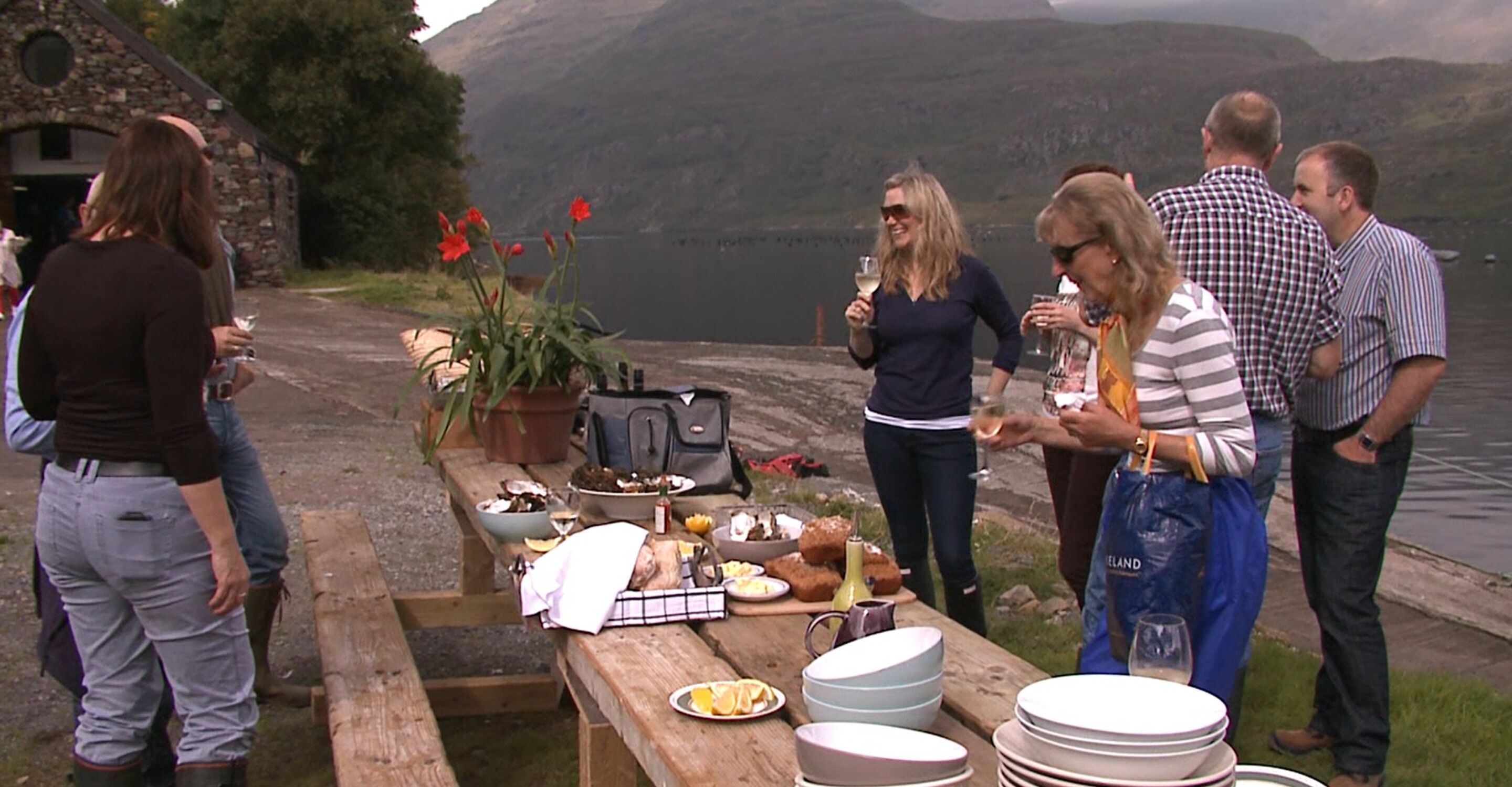 A group of people around a picnic table with food and drinks, overlooking a lake with mountains in the background.