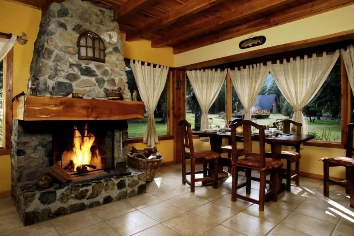 A cozy room with a stone fireplace, wooden ceiling, and a dining area with chairs and a table.