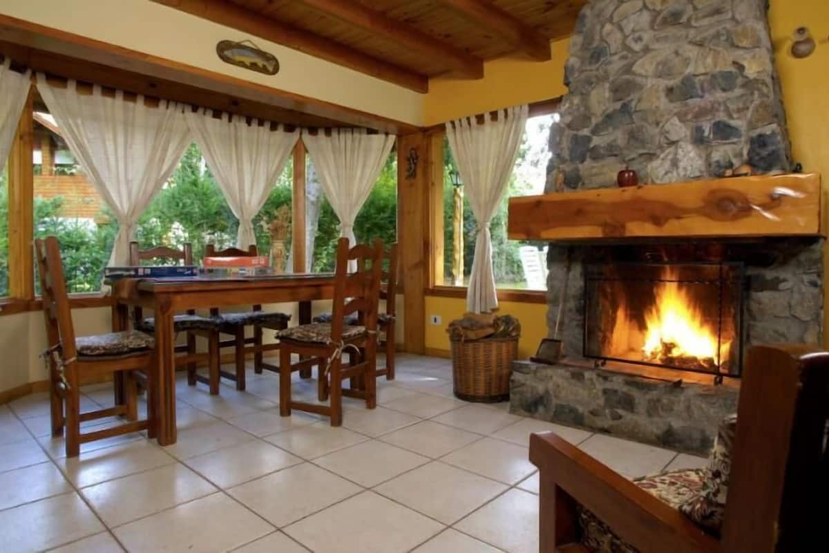 A cozy room with a stone fireplace, wooden furniture, and large windows with curtains.