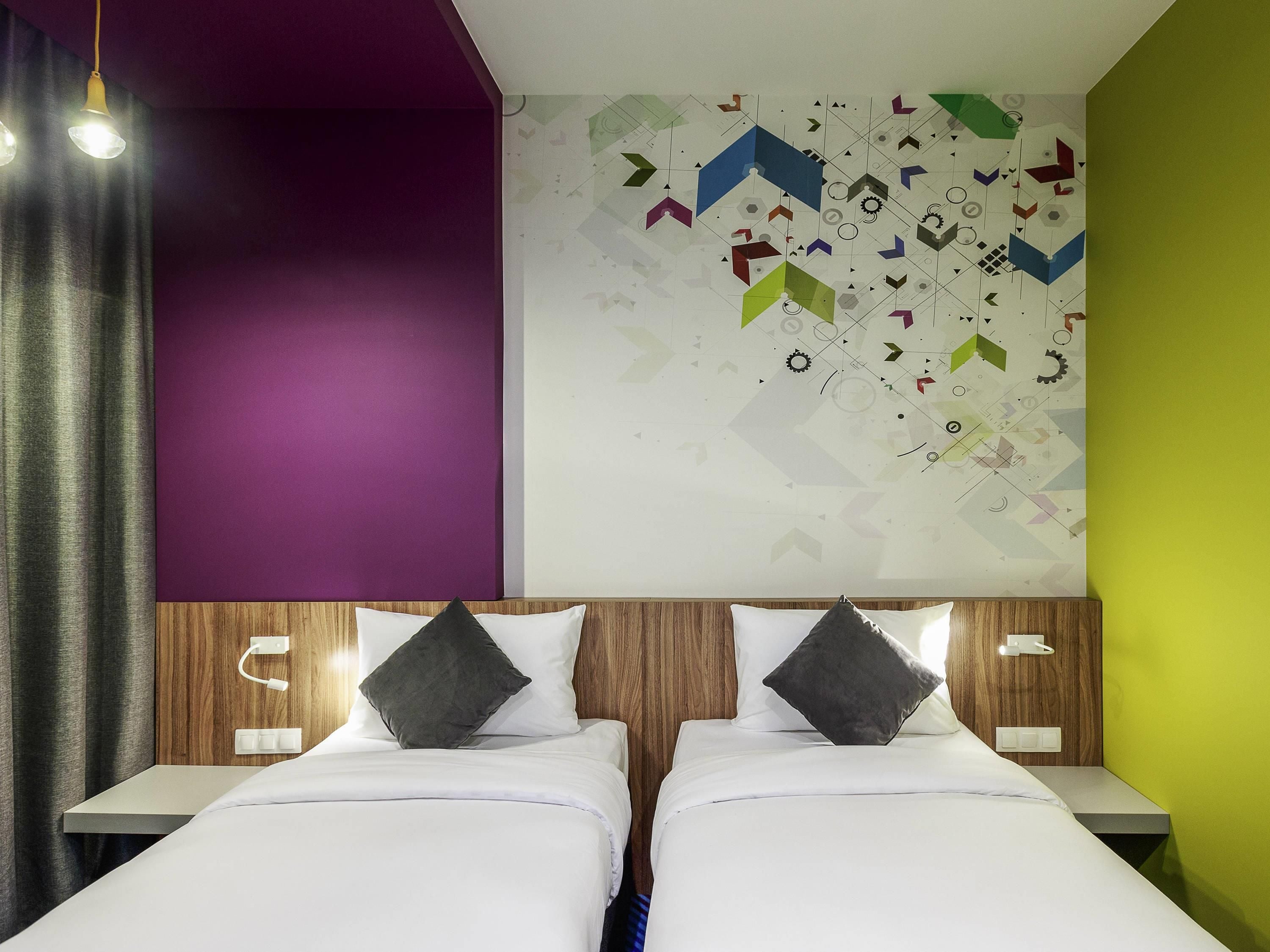 Ein modernes Hotelzimmer mit zwei Betten, einer lilafarbenen Akzentwand, einem geometrischen Wandbild und einem Holzkopfende.