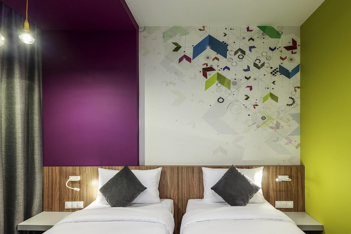 Una habitación de hotel moderna con dos camas, una pared de acento morada, un diseño artístico geométrico en la pared y un cabecero de madera.