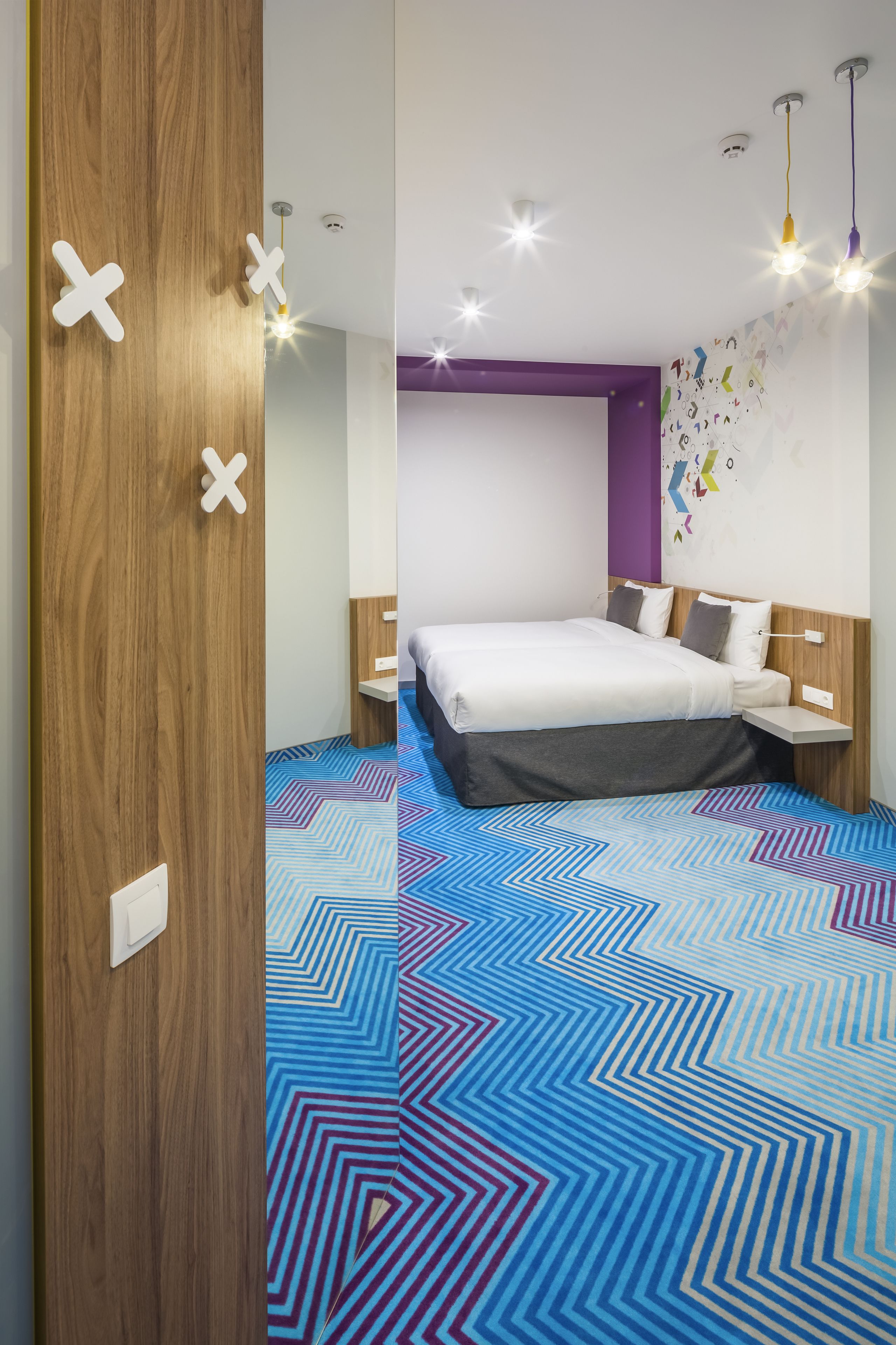 A modern hotel room with a large bed, a wooden door, and a colorful geometric carpet.