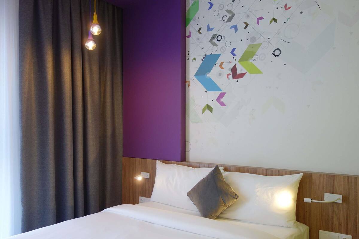 A hotel room with a bed, curtains, a wall with geometric patterns, and a light fixture.
