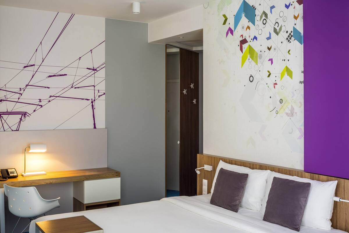 A hotel room with a bed, desk, chair, and a wall with geometric patterns.