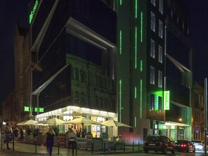 A modern building with illuminated vertical lines, outdoor seating with umbrellas, and a street with cars. - Ibis Styles Lviv Center (Lviv)