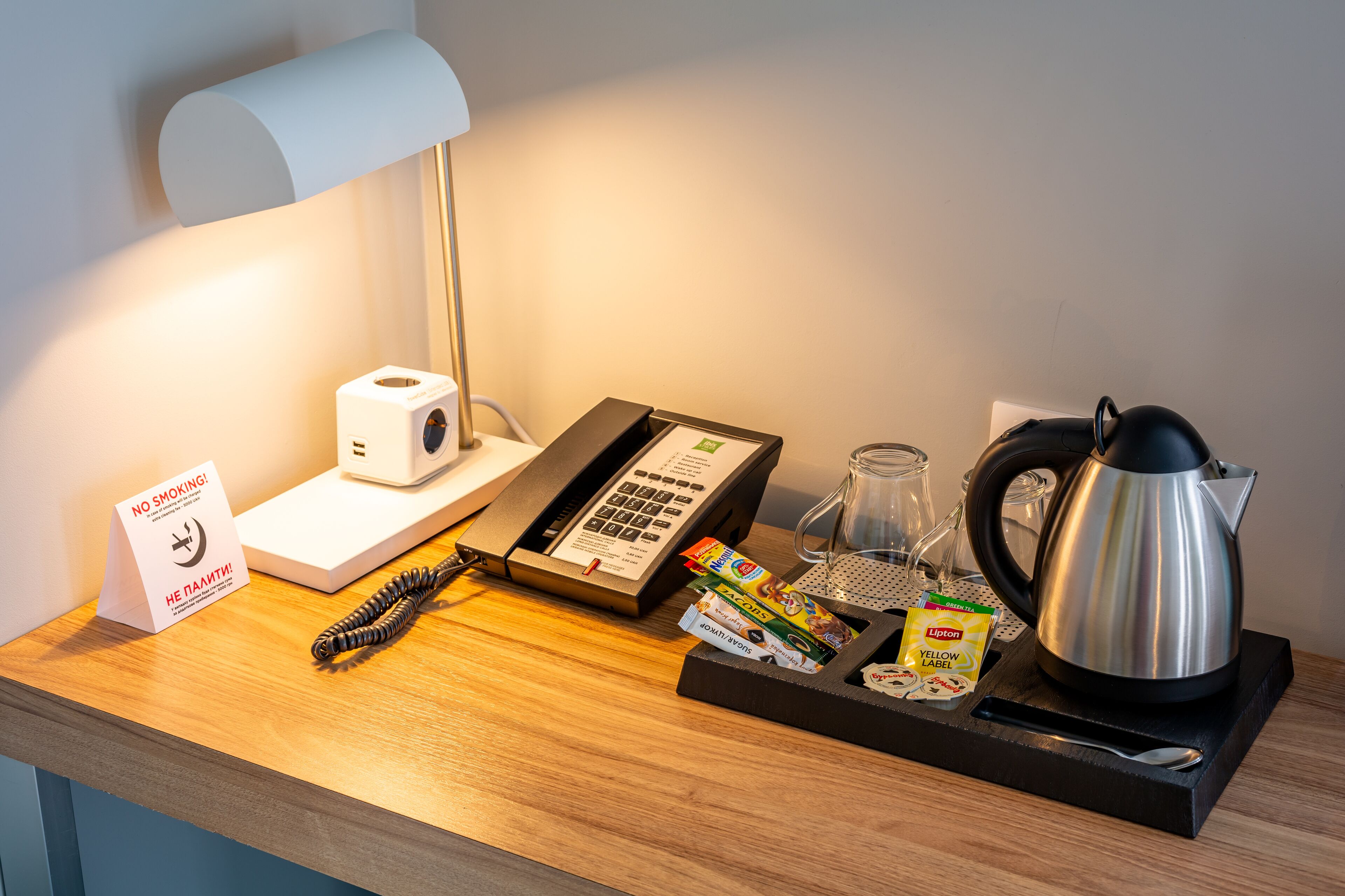 Mostrador de recepción de hotel con teléfono, cafetera, bandeja de snacks, hervidor de agua y cartel de 'No Fumar'.