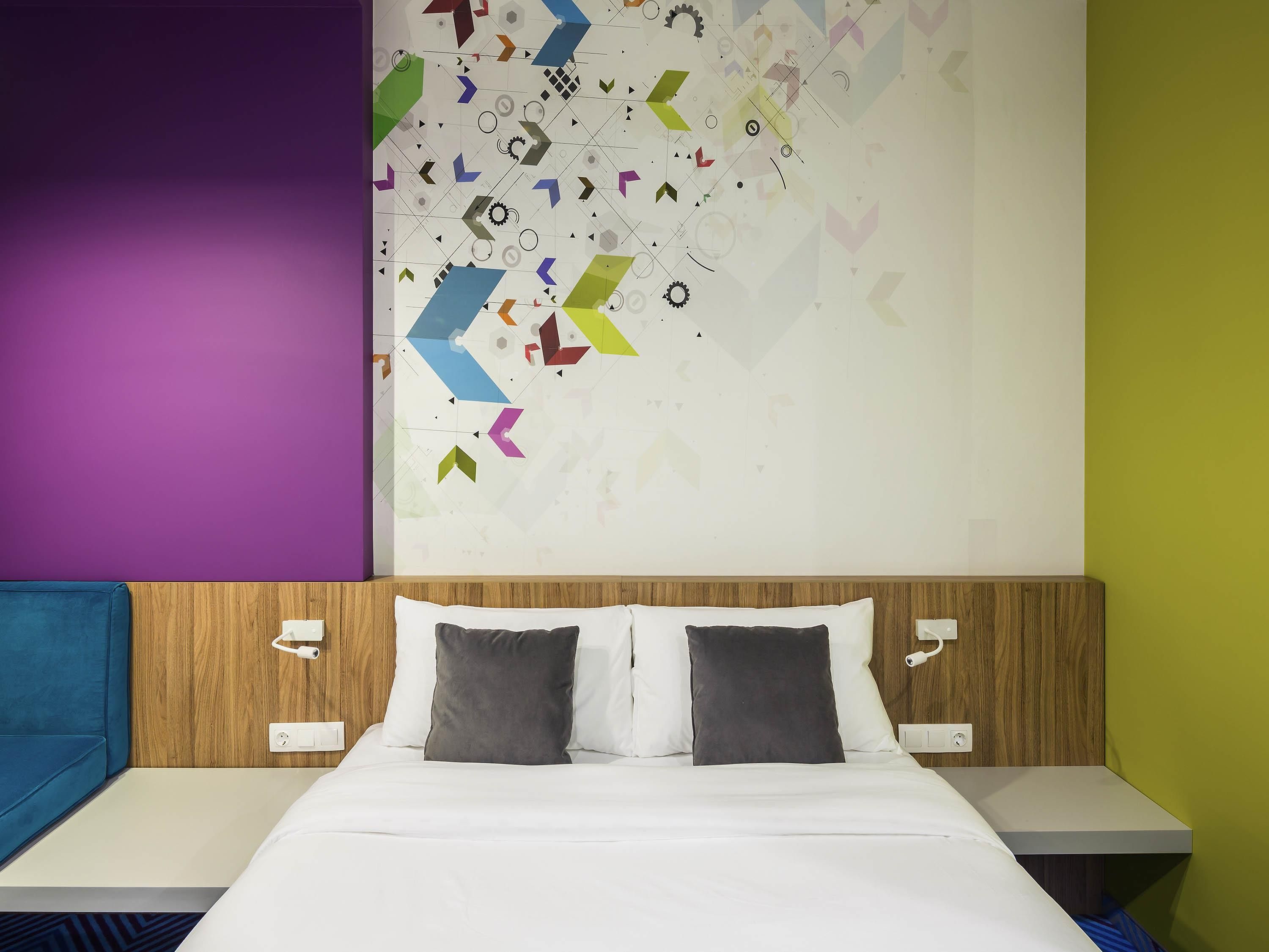Ein modernes Hotelzimmer mit einem Bett, zwei Kissen, einem Holzkopfende und einem farbenfrohen, abstrakten Wanddesign.