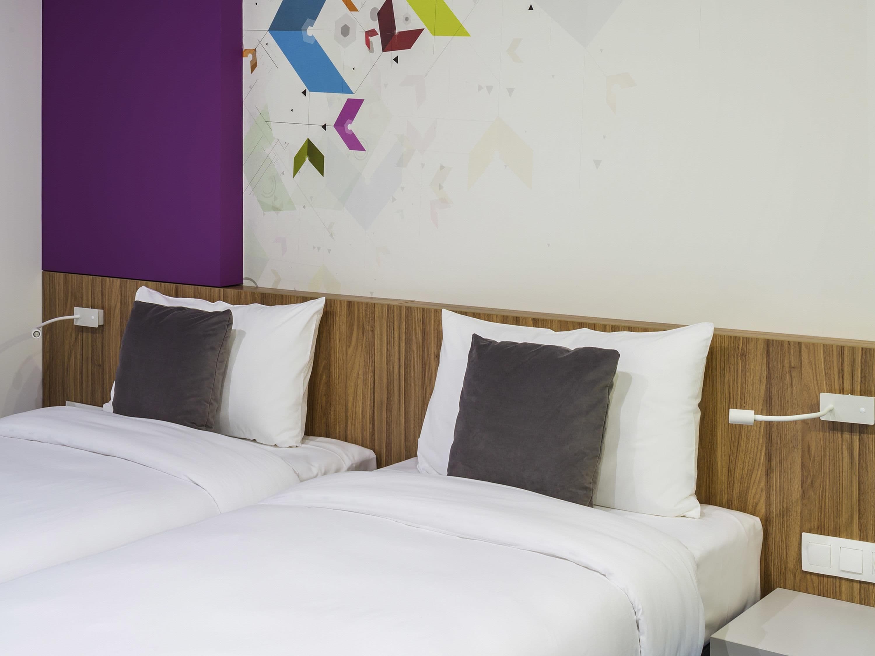 Habitación de hotel con cabecera de madera, dos camas con ropa blanca, una pared de acento morada y decoración geométrica en la pared.