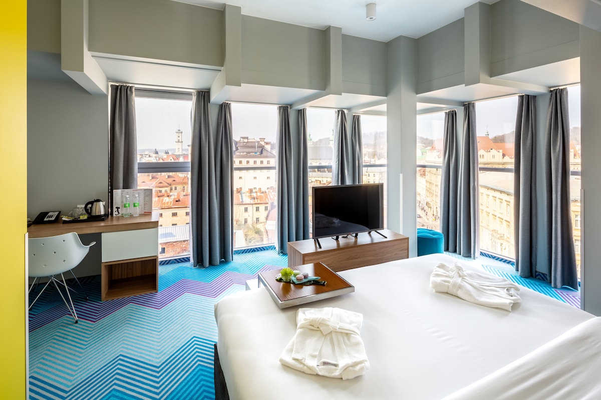 A hotel room with a large bed, a desk, a TV, and a view of the city.