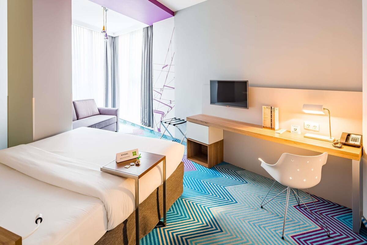 A hotel room with a bed, a desk with a computer, a TV, and a chair.