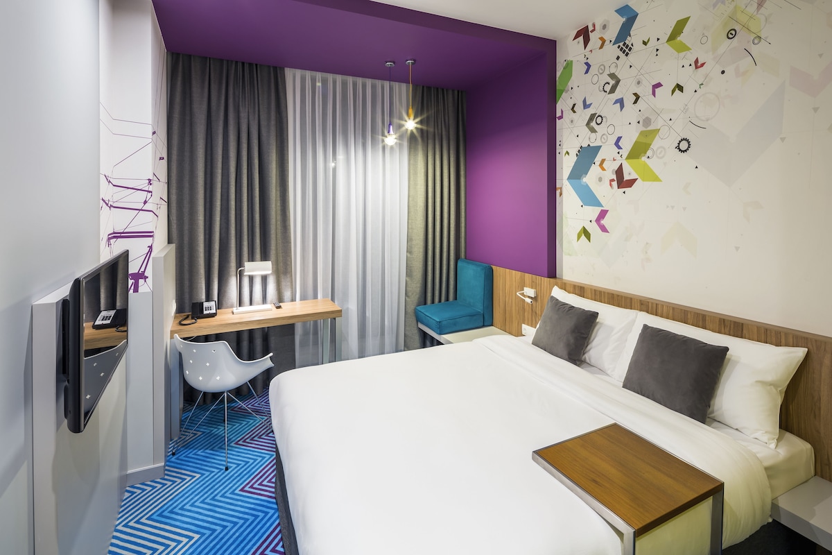A modern hotel room with a large bed, a desk, a chair, and a wall with geometric patterns.