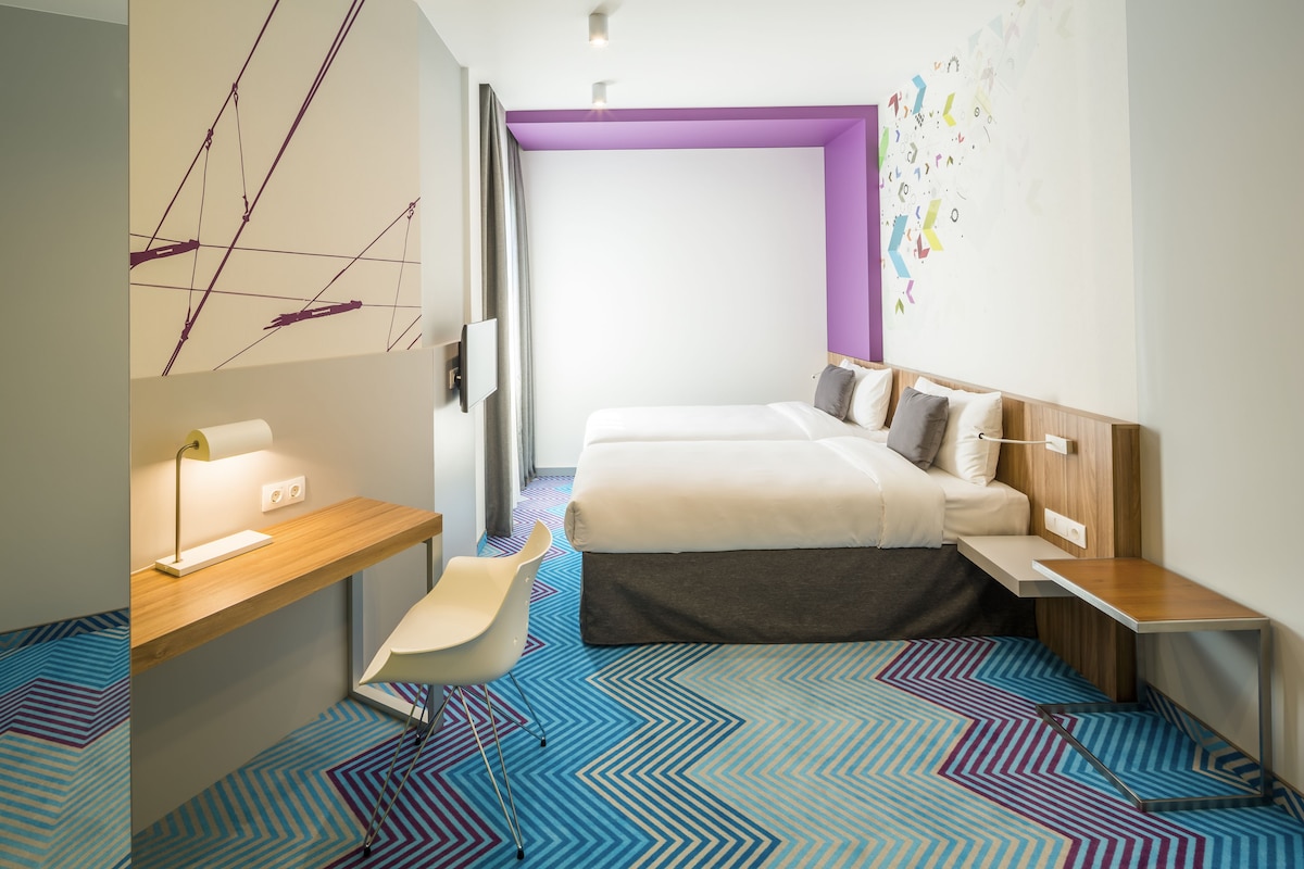 A modern hotel room with a bed, a desk, a chair, and a colorful geometric carpet.
