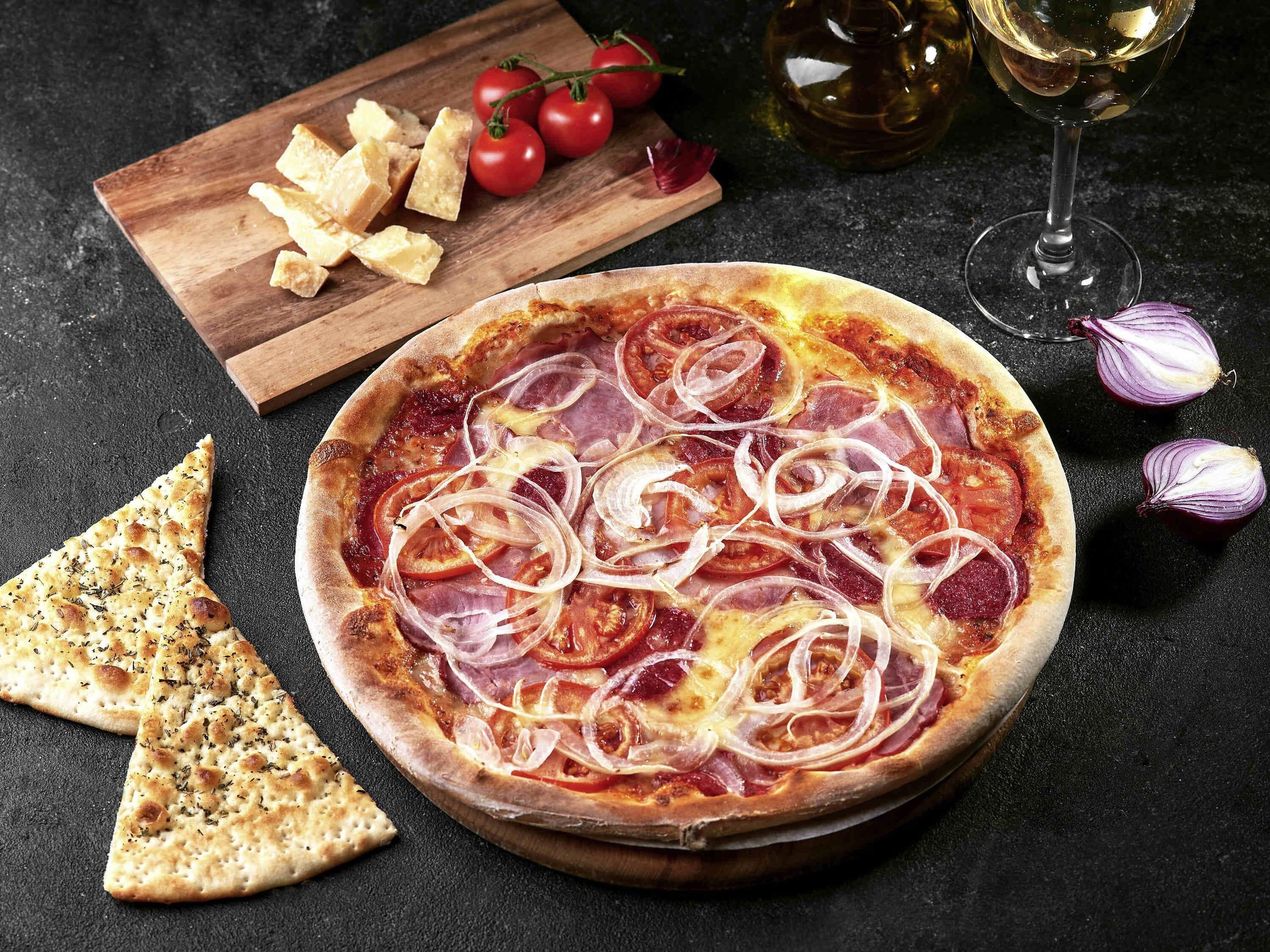A pizza with toppings, a wooden board with cheese and tomatoes, and a glass of white wine.