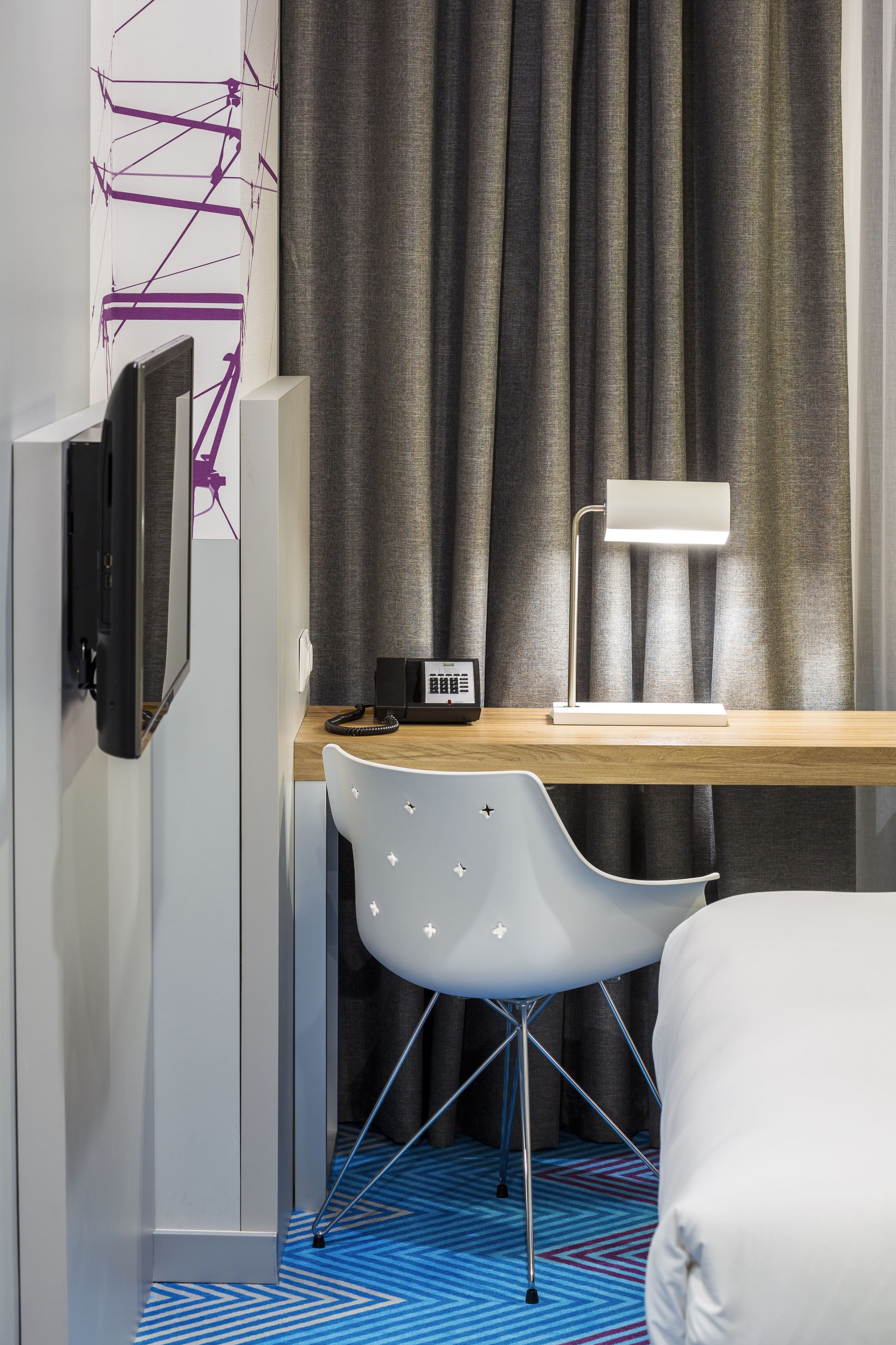 Ett modernt hotellrum med ett skrivbord i trä, en vit stol, en säng, en tv som är monterad på väggen och en skrivbordslampa.