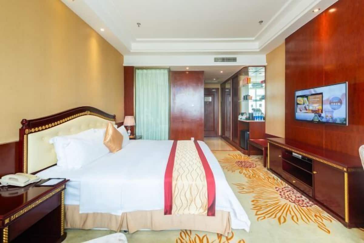 Quarto de hotel com uma cama grande, televisão, secretária e casa de banho visível através de uma porta aberta.