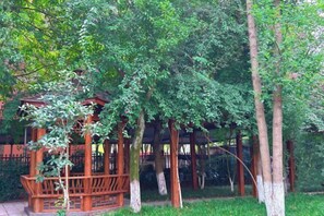 Un belvédère en bois, doté d’un toit en treillis, entouré d’une végétation luxuriante et de grands arbres.