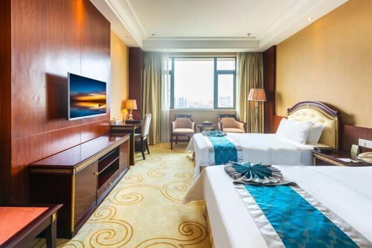 Quarto de hotel com duas camas, uma escrivaninha, televisão e janela com cortinas.