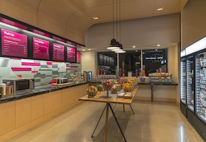 A modern cafe with a wooden counter, a menu board, and a refrigerator filled with beverages. - Aloft by Marriott College Station (College Station)