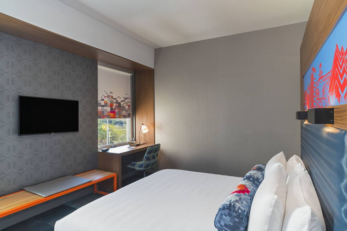 A modern hotel room with a large bed, a desk with a chair, a TV mounted on the wall, and a window with a view of greenery.