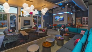 A modern lounge area with multiple seating options, including sofas and chairs, arranged around a central fireplace. There are several pendant lights hanging from the ceiling, and a flat-screen TV mounted on the wall. - Aloft by Marriott College Station (College Station)
