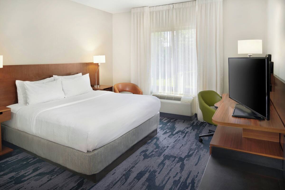 A hotel room with a large bed, a desk with a flat-screen TV, a chair, and a window with curtains.